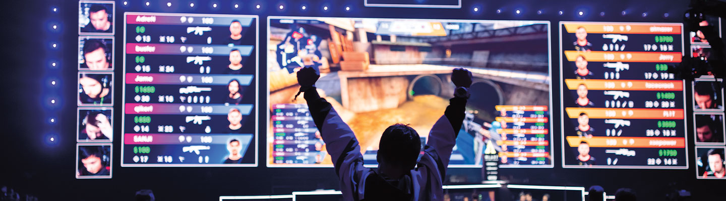 A person raising both arms in victory in front of a huge screen showing a virtual arena