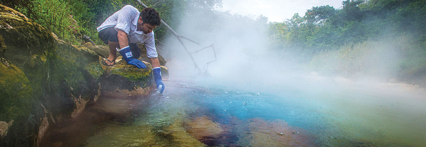 A man with gloves on collecting samples from a boiling river 