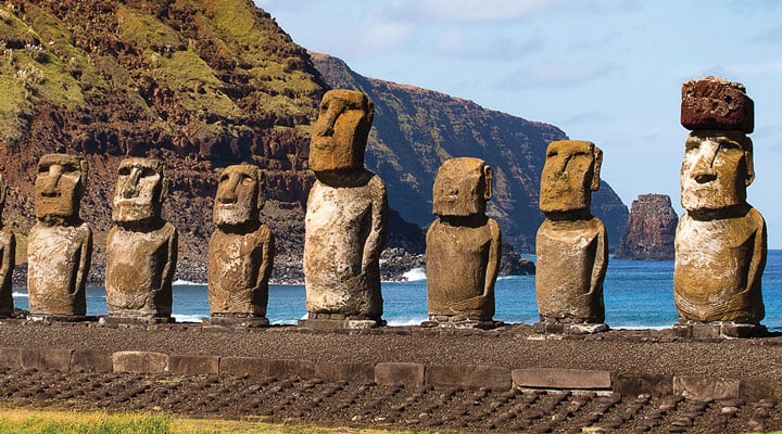 Seven moai statues stand in a row on a stone platform with ocean and mountains behind them