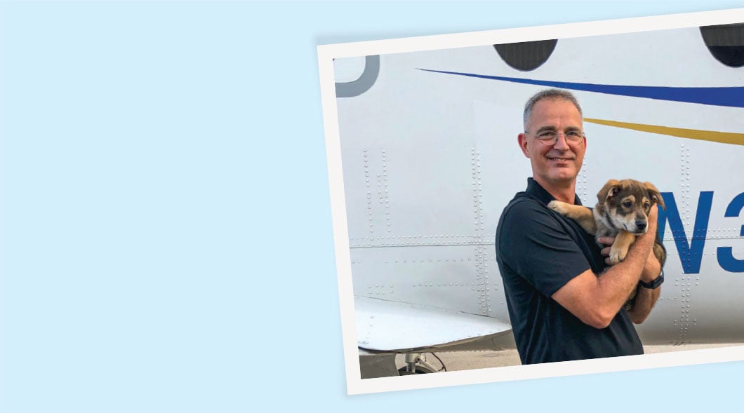 A man holds a small puppy in front of a small aircraft