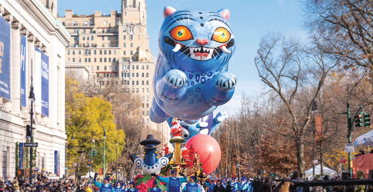 A giant blue cat balloon floats above a parade float while handlers in blue walk the crowded street